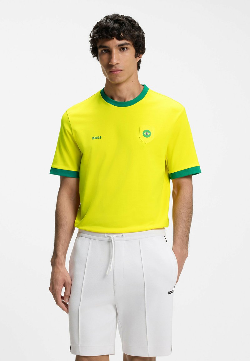 Jeune homme portant un t-shirt jaune avec un liseré vert et un petit emblème du drapeau brésilien, assorti à un short blanc à cordon.