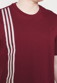 Burgundské tričko vyrobené z bavlny, s šedými svislými pruhy na levé straně a malým logem na hrudi.