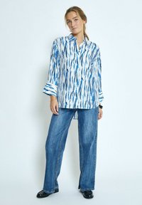 Camicia a maniche lunghe con bottoni di colore bianco con motivi astratti blu, abbinata a jeans a vita alta e gamba larga. Le scarpe nere completano l'outfit.