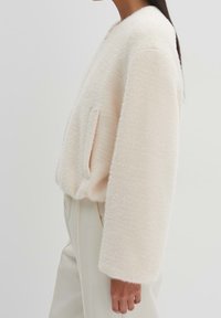 Kort, krämfärgad cardigan i en texturerad, luddig tyg, med vida ärmar och ett enda sidofack. Minimalistisk design utan knäppningar.