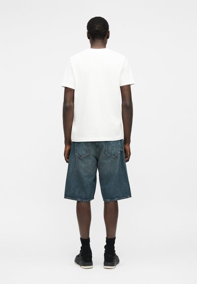 Άνδρας που στέκεται με την πλάτη γυρισμένη, φοράει απλό λευκό κοντομάνικο πουκάμισο, τζιν σορτς μέχρι τα γόνατα, μαύρες κάλτσες και μαύρα αθλητικά παπούτσια.