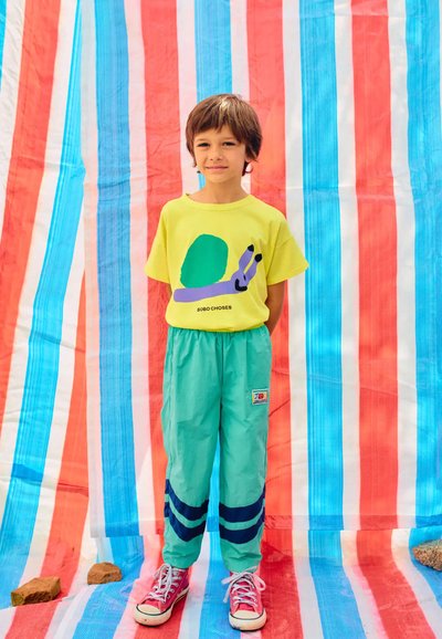 Helles gelbes T-shirt mit Grafikdesign, hellgrüne Hose mit marineblauen Streifen und rote Turnschuhe. Der Hintergrund zeigt ein bunt gestreiftes Stoffmuster.