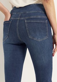 Mörkblå denimjeans med en figurnära design, som har två bakfickor, synliga sömmar och ett något strukturerat material.