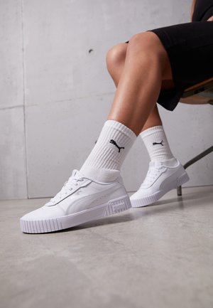 Piernas con zapatillas blancas Puma y calcetines blancos Puma, cruzadas en los tobillos, sentadas en una silla frente a una pared gris.
