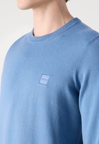 Pull en tricot bleu clair à col rond, avec un petit patch carré portant le logo "BOSS" sur la poitrine. Texture douce, design simple.