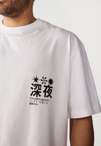 Camiseta de algodón blanca con un estampado gráfico negro que incluye caracteres japoneses y formas abstractas en el área del pecho izquierdo. Mangas cortas.