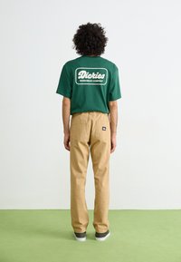 Grøn bomulds t-shirt med hvid "Dickies Workwear Company" logo på ryggen, kombineret med beige lige ben bukser og grå sneakers.