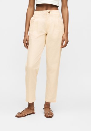 Kvinde iført beige bukser med høj talje med knaplukning og brune flade sandaler med stropper, stående mod en ensfarvet baggrund.