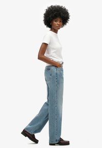 Jasnoniebieskie spodnie typu wide-leg z wyblakłym wykończeniem, w połączeniu z gładką białą koszulką. Model nosi ciemne, błyszczące buty. Prosty, casualowy styl.