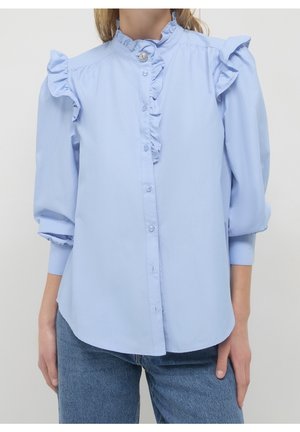 Femme portant un chemisier bleu clair avec des épaules et un col à volants, des manches trois-quarts bouffantes, et un devant boutonné, associé à un jean bleu.