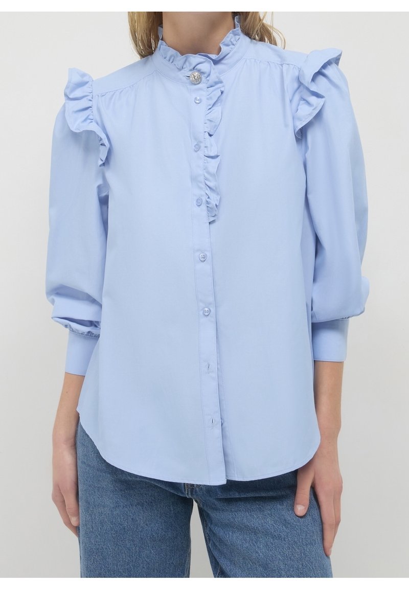Femme portant un chemisier bleu clair avec des épaules et un col à volants, des manches trois-quarts bouffantes, et un devant boutonné, associé à un jean bleu.