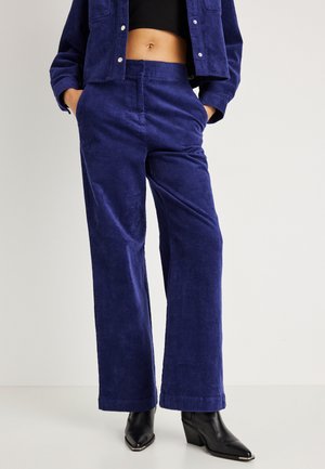 Nainen, jolla on ylhäältä vyötäisille ulottuvat, leveälahkeiset siniset corduroypöksyt, niihin sopiva lyhyt takki, musta lyhyt toppi ja mustat kärkitossut.