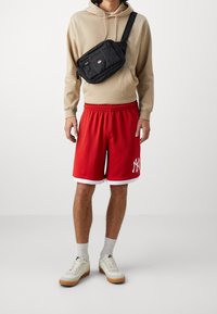 Shorts de basketball rouges avec bordure blanche et logo NY, sweat à capuche beige, sac bandoulière noir, chaussettes gris clair et baskets crème avec semelles en gomme.