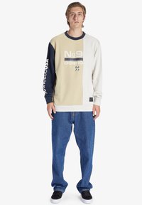 Sudadera de gran tamaño en beige, gris y marino; presenta un estampado gráfico y puños acanalados, combinada con jeans sueltos azules y zapatillas negras.
