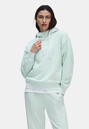 Femme aux cheveux foncés portant un sweat à capuche vert menthe clair et un pantalon assorti, posant avec une main près du col.