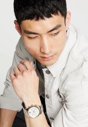 Silver and gold-tone wristwatch with a white dial, Roman numerals, chronograph features, and a patterned metal band.