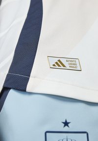 Jersey atlético azul claro con un patrón texturizado. Presenta detalles en azul marino y una etiqueta de licencia auténtica en dorado y blanco.