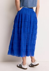 Femme portant une jupe midi plissée bleu royal à couches, un gilet en denim court et des baskets blanches Adidas avec des rayures noires, adossée à un mur.