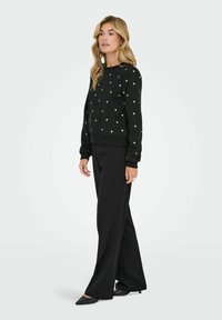 Zwarte sweatshirt met gouden hartpatronen, ronde hals en geribde manchetten, gecombineerd met zwarte wijde broek en puntige zwarte schoenen.