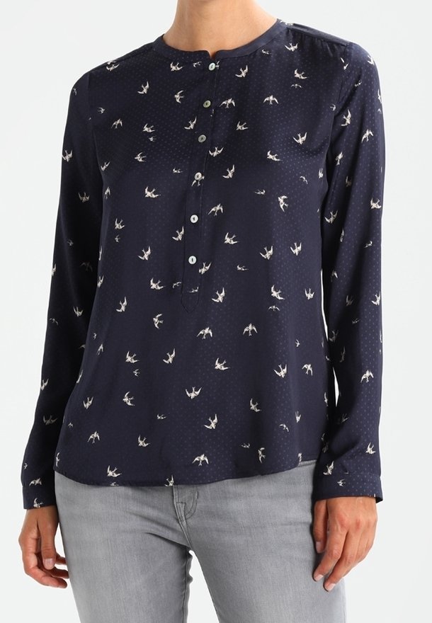 Blouse à manches longues bleu marine avec petits imprimés d'oiseaux blancs et patte de boutonnage, portée avec un pantalon gris clair.