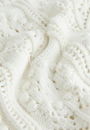 Weiße gestrickte Decke mit einem strukturierten Design, das aus filigranen Mustern und offenen Maschen besteht und ein warmes, gemütliches Erscheinungsbild schafft.