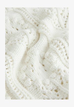 Balta adīta sega ar teksturētu dizainu, kurā ir izsmalcinātas rakstu detaļas un atvērtas cilpas, radot siltu un mājīgu izskatu.