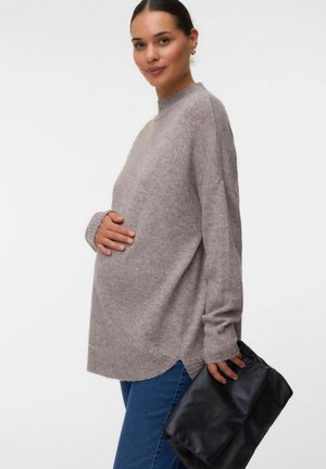 Femme enceinte portant un pull gris ample et un jean, tenant une pochette noire, caressant doucement son ventre et souriant doucement.