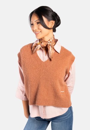 Kvinne smilende, iført en lyserosa skjorte, brun strikket vest, brunt mønstret hals skjerf og blå jeans mot en ensfarget bakgrunn.