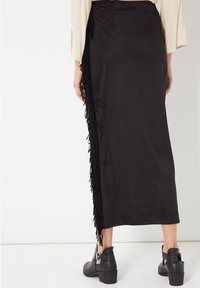 Falda maxi de ante negro con detalle de flecos en el lateral. Textura suave, diseño recto y combinada con botines de tobillo con hebillas decorativas.