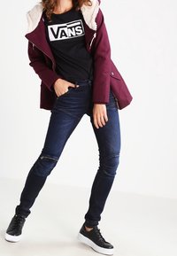 Vinrød jakke med hætte over en sort Vans t-shirt, kombineret med mørkeblå skinny jeans med lynlåselementer og sorte sneakers.