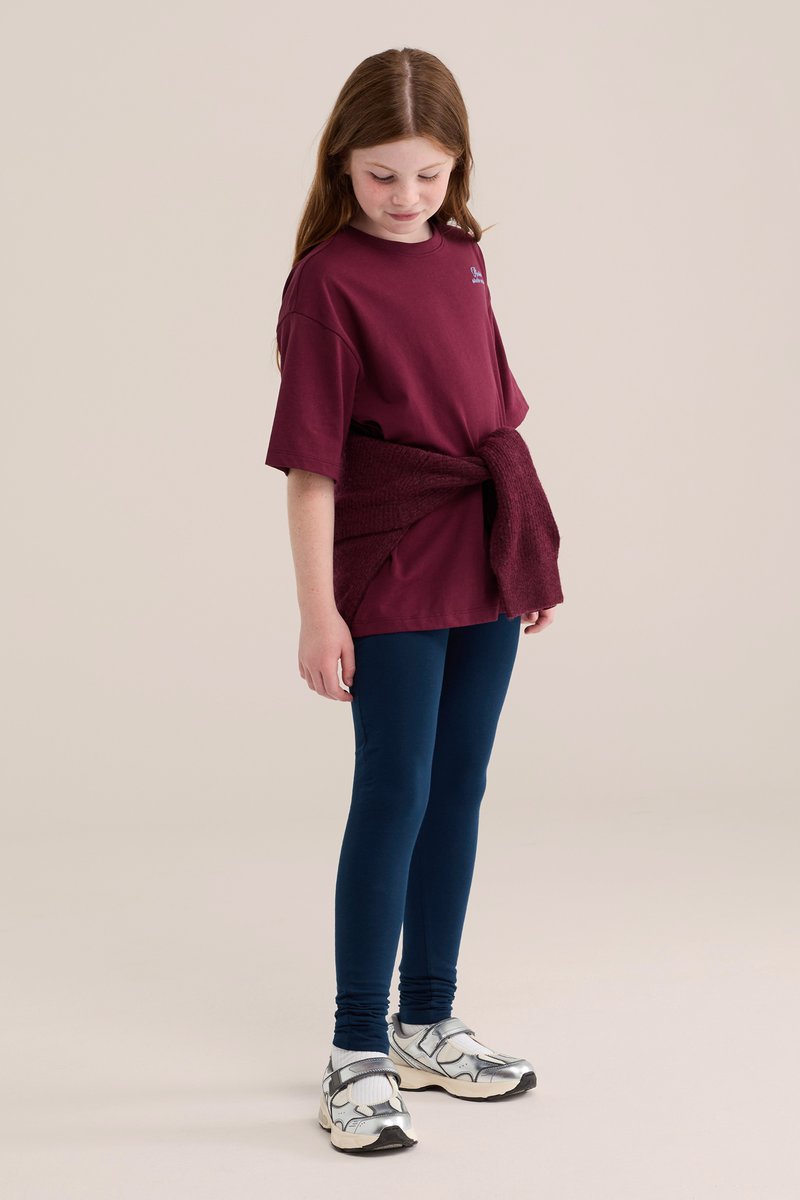 Ragazza giovane che indossa una t-shirt color amaranto con un maglione abbinato legato in vita, leggings blu, calzini bianchi e scarpe da ginnastica argento.