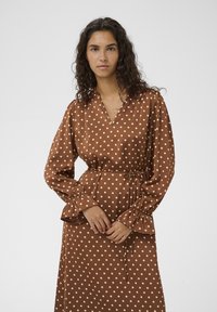 Rjava obleka z belimi pikami, V-izrezom, pasom in dolgimi, razširjenimi rokavi. Gladek material s kremasto obarvanimi pikami.