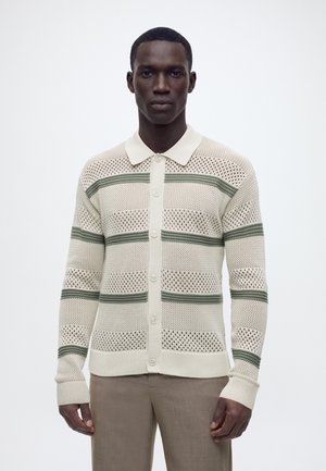 Mann trägt beigen Strickcardigan mit Knöpfen und grünen horizontalen Streifen sowie beige Hose, steht vor einem schlichten weißen Hintergrund.