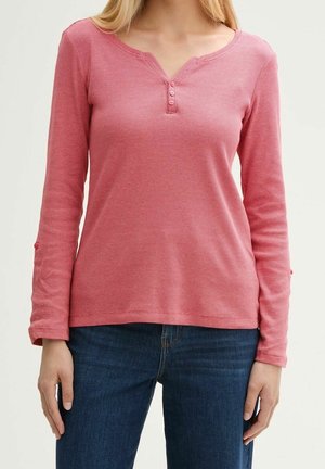 Kobieta ubrana w różową, prążkowaną koszulkę henley z długim rękawem i trzema guzikami oraz niebieskie dżinsy, stojąca na jednolitym tle.