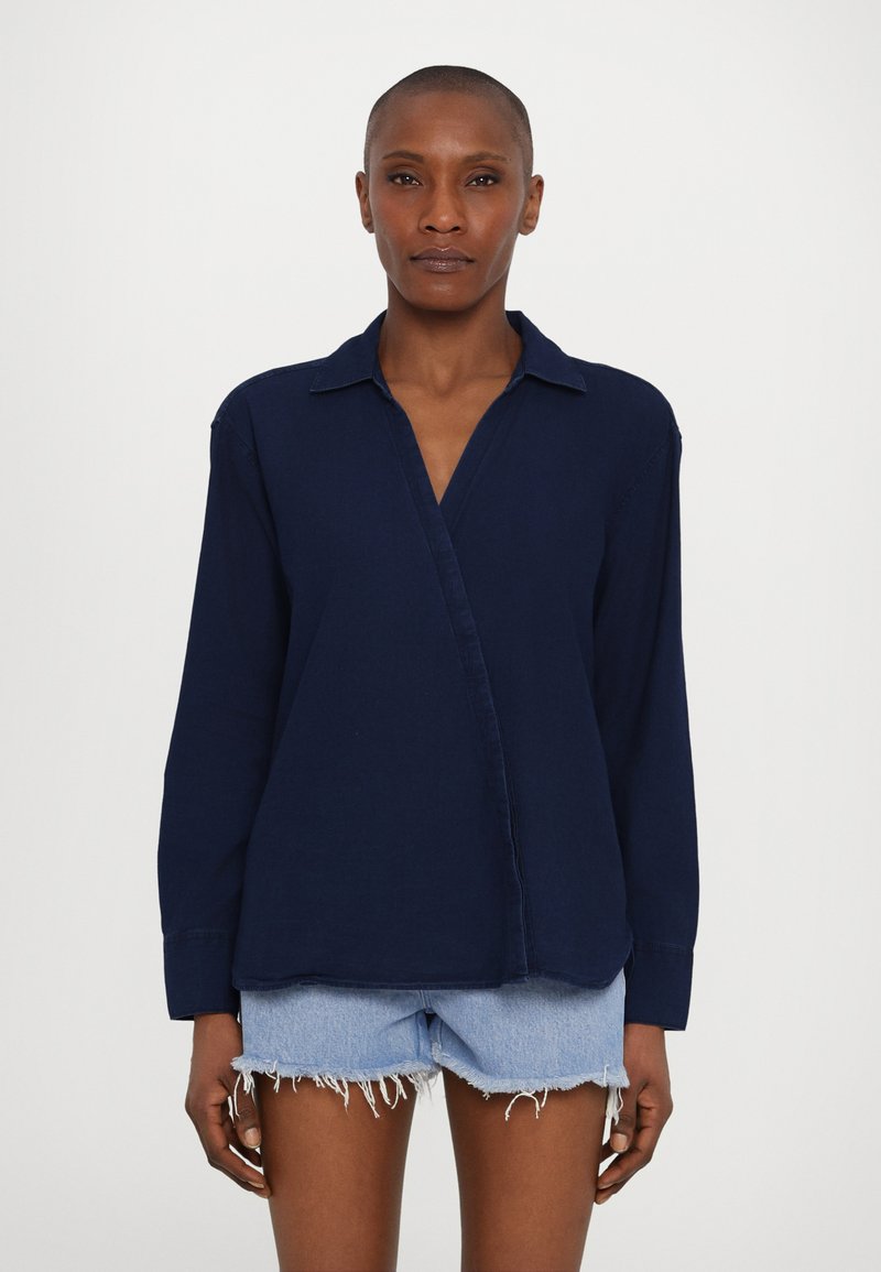 Mörkblå långärmad skjorta med krage, med en diagonalt framfälld placket och avslappnad passform. Bärs med fransiga denimshorts.