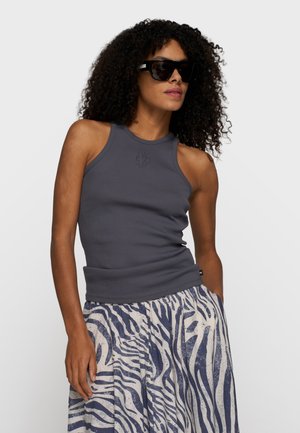 Donna con capelli ricci che indossa occhiali da sole neri, un top aderente senza maniche grigio scuro e una gonna a vita alta con strisce zebrate blu e bianche.