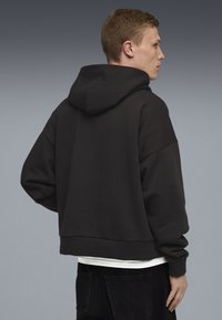 Ung mand set fra siden iført en sort oversized hættetrøje over en hvid skjorte og sorte bukser mod en grå baggrund.