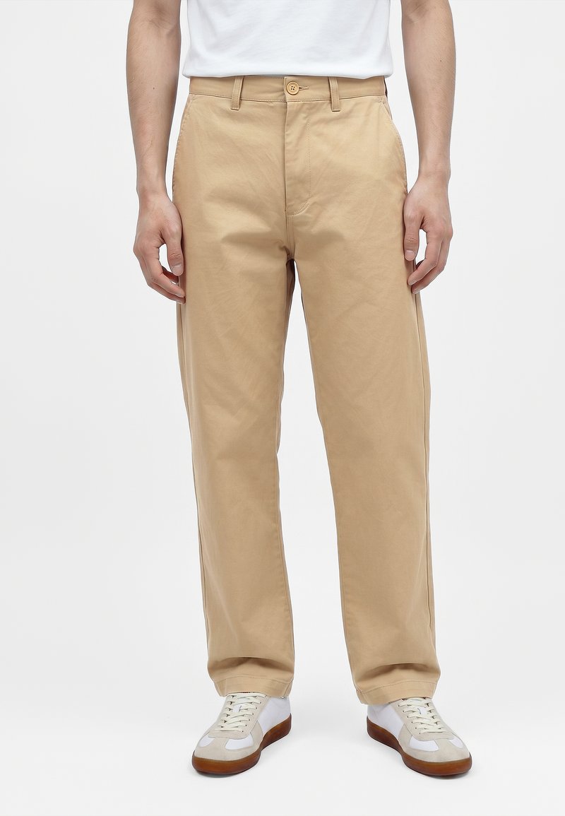 Homme portant un pantalon beige à jambes droites, des baskets blanches avec des semelles marron et un t-shirt blanc, debout devant un fond uni.