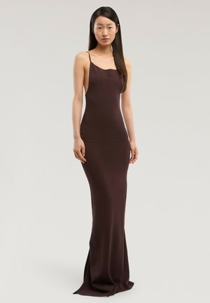 Mujer con cabello largo y negro, vestida con un vestido de noche sin mangas ajustado de color marrón oscuro, posando frente a un fondo claro y sencillo.