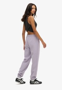 Grijze cropped tanktop en lila sweatpants met elastische boorden. Model draagt zwarte sportschoenen. Neutrale achtergrond.