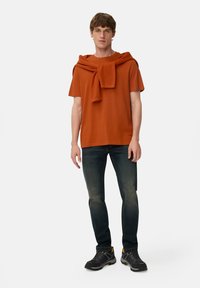 Rustrød t-shirt med rund hals, parret med mørk denim jeans. En matchende rustrød trøje hængt over skuldrene. Sorte sko.