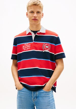 Joven con las manos en los bolsillos que lleva una camiseta tipo polo a rayas rojas, azul marino y blancas con nombres de ciudades y bordado de logotipo, y vaqueros azul claro.