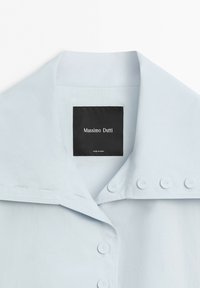 Colletto di camicia azzurro chiaro con quattro bottoni sul lato destro e un'etichetta nera con la scritta Massimo Dutti cucita all'interno della scollatura.
