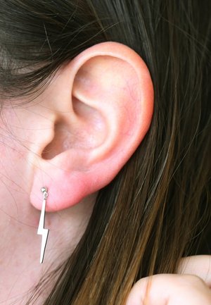 Zilveren oorring in de vorm van een bliksemflits met een klein bolletje als bevestiging, weergegeven tegen een achtergrond van donker haar en huid.
