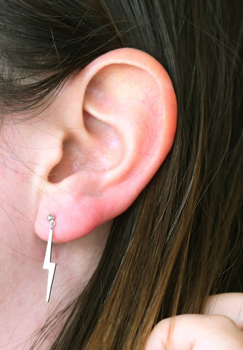 Silberne Ohrringe in Form eines Blitzes mit einem kleinen Kugelstecker, präsentiert vor einem Hintergrund aus dunklem Haar und Haut.