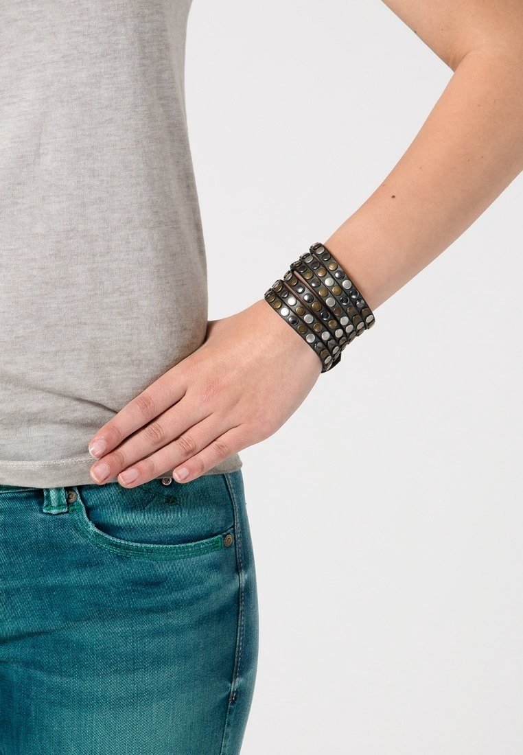 Bras droit plié avec la main reposant sur la hanche, portant un bracelet enveloppant gris clouté, une chemise grise et un jean bleu sur un fond clair.