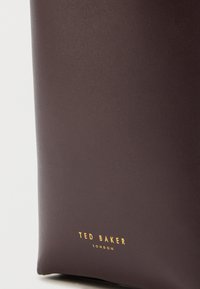 Part of a dark brown leather bag showing embossed gold "Ted Baker London" logo near the bottom edge.
