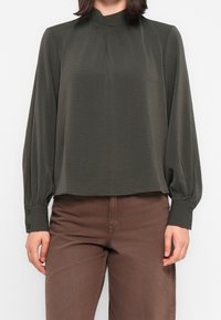Blouse vert foncé avec un col montant, un détail plissé et de longues manches bouffantes. Les poignets sont ornés de boutons, et le tissu semble doux et léger.