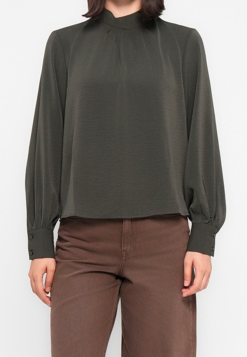 Blouse vert foncé avec un col montant, un détail plissé et de longues manches bouffantes. Les poignets sont ornés de boutons, et le tissu semble doux et léger.