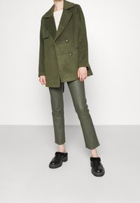 Persoon in een olijfgroene dubbelzijdige wollen jas, olijfgroene leren broek en zwarte instappers met bontafwerking, staand op een grijze vloer.